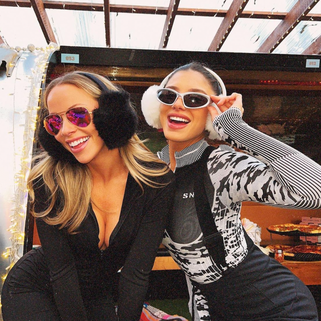 Two women posing together outdoors, one wearing sunglasses and the other with earmuffs.