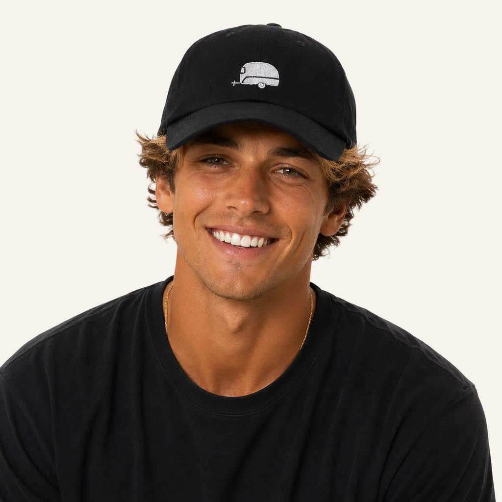 A smiling young man wearing a black baseball cap with a small white graphic logo and a black t-shirt, photographed against a solid white background.