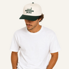 A man wearing a cream and dark green baseball cap with 'BROKENDOWN PALACE' and a cigarette graphic printed on the front, paired with a plain white t-shirt against a solid white background.