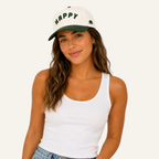 A woman wearing a white baseball cap with a dark green brim and 'HAPPY' embroidered in green on the front, paired with a white ribbed tank top and blue jeans, against a solid white background.