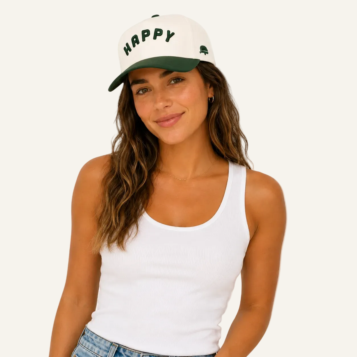 A woman wearing a white baseball cap with a dark green brim and 'HAPPY' embroidered in green on the front, paired with a white ribbed tank top and blue jeans, against a solid white background.