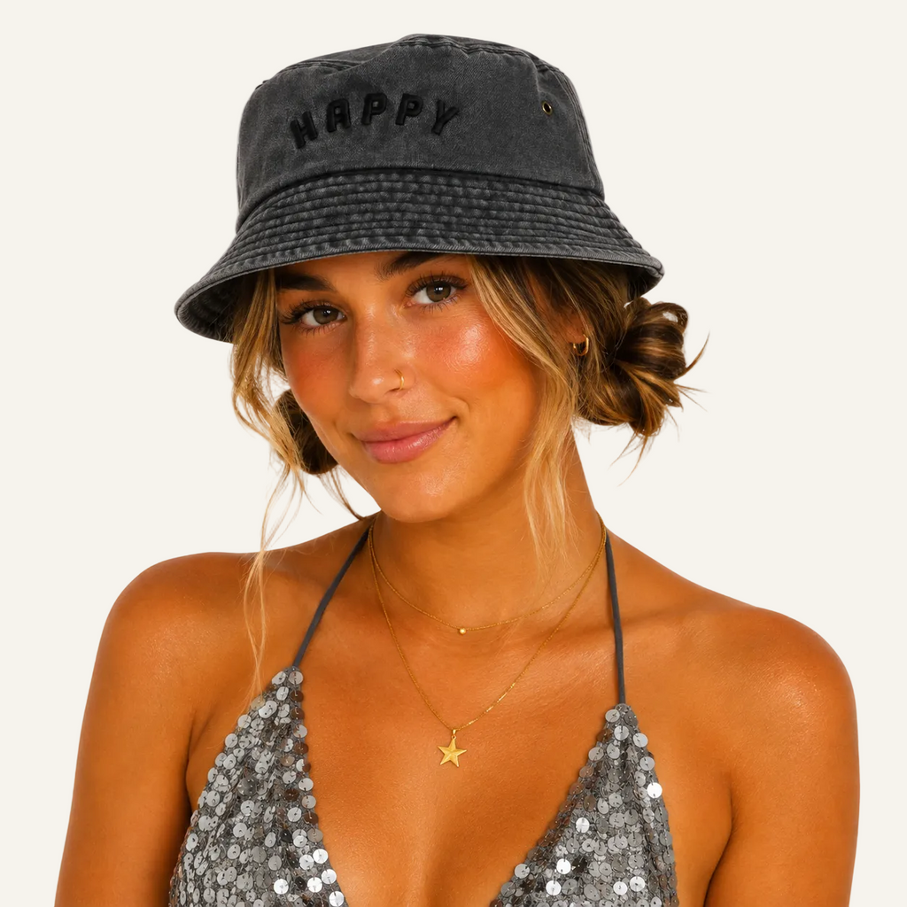 A woman wearing a dark gray washed bucket hat with the word 'HAPPY' embroidered on the front, paired with a silver sequined halter top against a plain white background.
