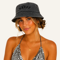 A woman wearing a dark gray washed bucket hat with the word 'HAPPY' embroidered on the front, paired with a silver sequined halter top against a plain white background.