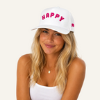 A smiling blonde woman wearing a white baseball cap with 'HAPPY' embroidered in pink across the front and a small pink logo on the side, against a plain white background.