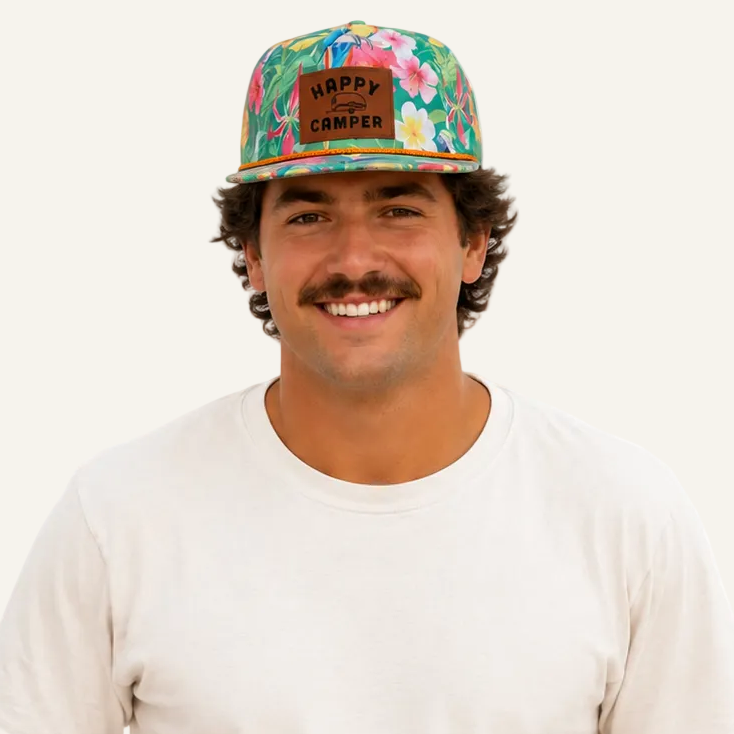 A smiling man wearing a colorful tropical print baseball cap with a brown patch that reads 'HAPPY CARNIVAL', a plain white t-shirt, and blue jeans, set against a solid white background.