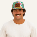 A smiling man wearing a colorful tropical print baseball cap with a brown patch that reads 'HAPPY CARNIVAL', a plain white t-shirt, and blue jeans, set against a solid white background.