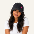 A woman wearing a black baseball cap with a small pink flamingo logo on the side, paired with a white t-shirt, photographed against a clean white background.
