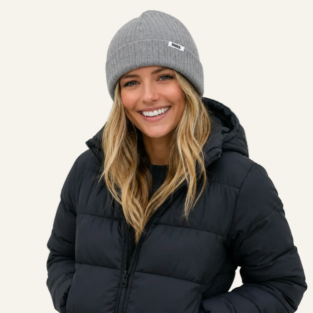 A smiling woman wearing a gray ribbed knit beanie with a small white logo patch and a black puffer jacket, photographed against a plain light background.