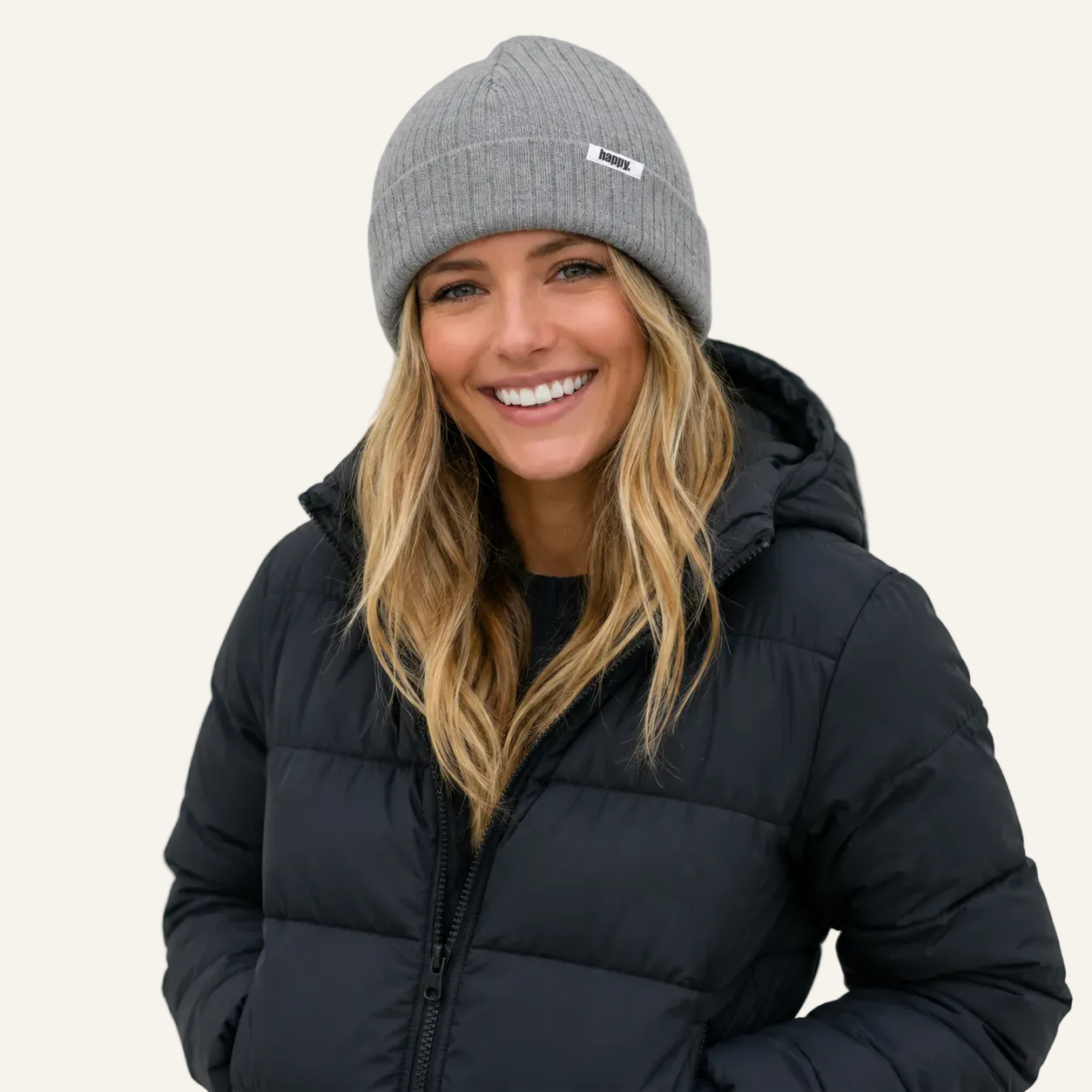 A smiling woman wearing a gray ribbed knit beanie with a small white logo patch and a black puffer jacket, photographed against a plain light background.